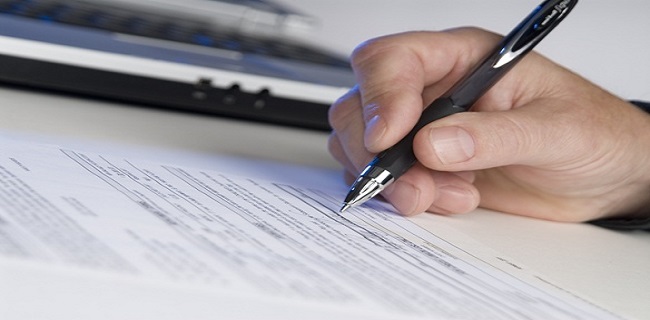man's hand holding a pen writing a signature, computer in background